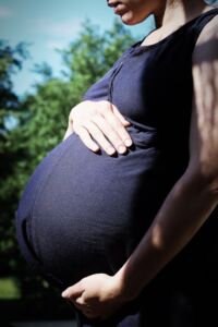 A pregnant woman wearing a fitted black dress gently hugging her belly with both hands. She stands in a soft-lit environment, her expression serene and loving, emphasizing the connection with her growing baby.