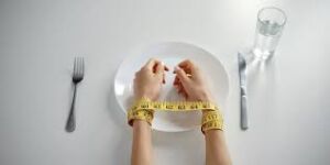 Eating Disorders Awareness: Symbolic image of a person sitting at a table with hands on an empty plate, tied with a measuring tape, depicting the challenges and complexities of the disorder.