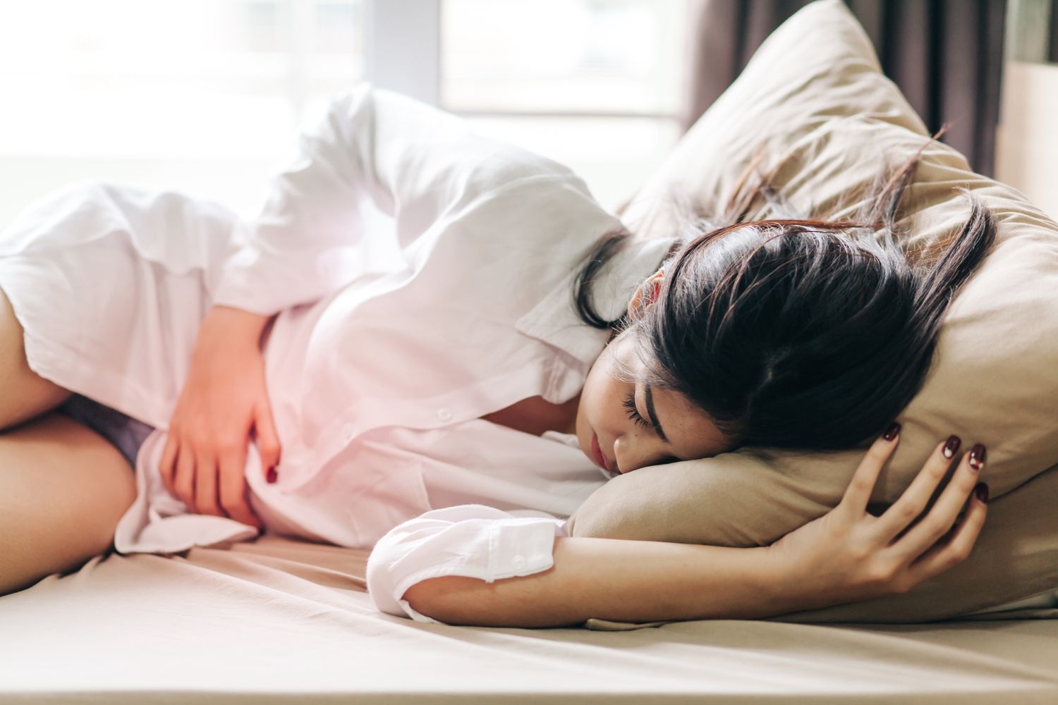 A woman lying in bed, holding her abdomen with a pained expression on her face. She is curled slightly to one side, suggesting discomfort, and her hand is gently pressing on her stomach. The bedding is soft and neutral, and the room appears calm, emphasizing her need for rest. The image conveys a sense of distress and discomfort, possibly related to menstrual cramps or digestive issues.