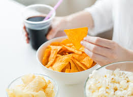 Person indulging in unhealthy snacks - An image showing a person eating a bowl of chips, popcorn, and a glass of juice, highlighting the consumption of food high in sodium, fats, and sugars.