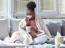 Woman with a common cold sitting on the couch sneezing in a tissue while wrapped in a blanket