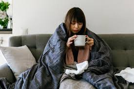 A sick individual sits on the sofa, wrapped in a cozy blanket, while sipping tea to soothe their ailment.
