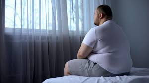 Overweight individual seated on bed, deep in thought, reflecting on the challenges and perceptions associated with obesity